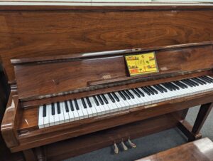 A polished walnut studio upright piano with the name Cora on it.