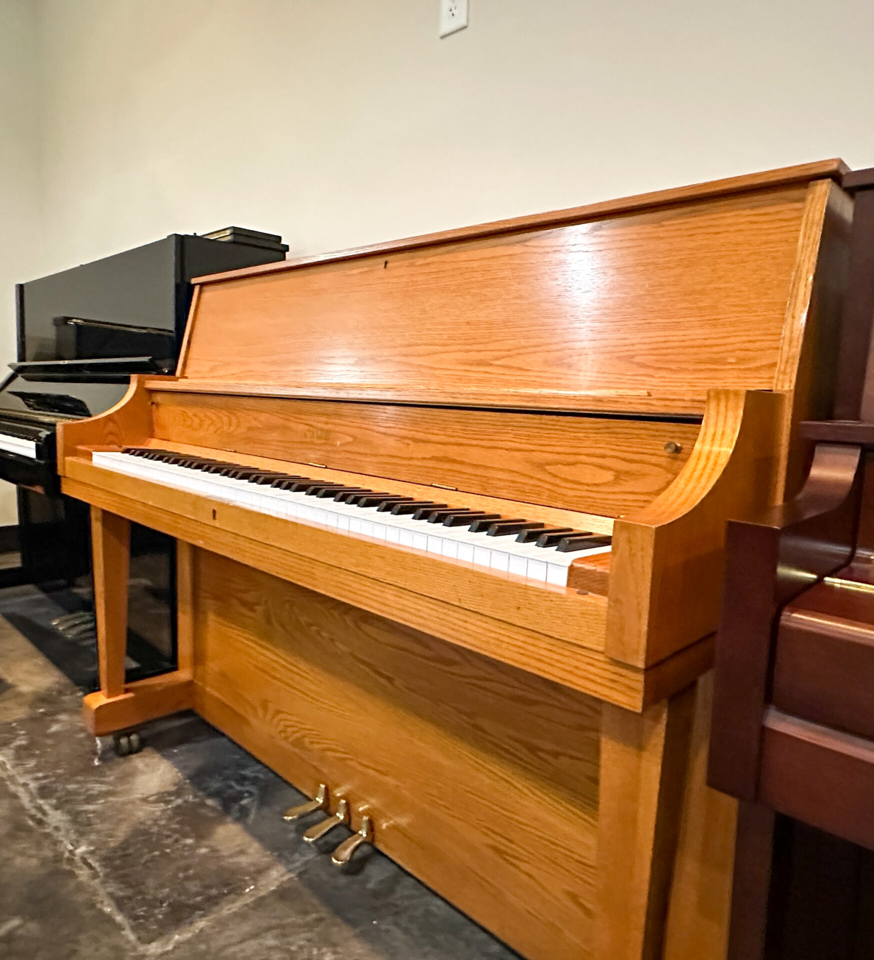 Side-angle view of a Boston UP-118 upright piano in oak finish, showing its elegant structure and three-pedal system.