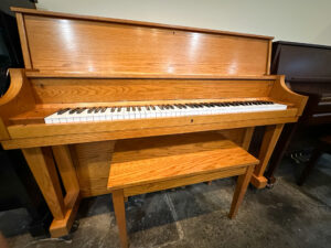 Used Boston UP-118 upright piano in oak finish with matching bench, displayed in a showroom setting.