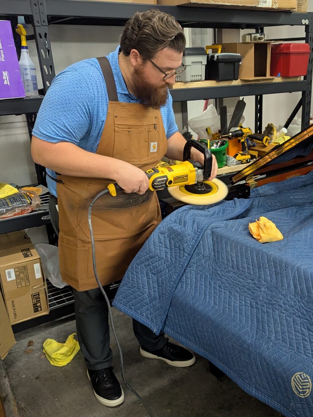 Forrest Mitchell carefully refinishing a grand piano in the workshop, using a professional buffing tool to restore its surface. Wearing a work apron, he demonstrates his craftsmanship and attention to detail in piano restoration.