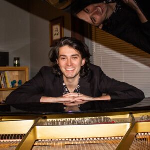 Piano teacher Nick Ricks, smiling at the camera with his arms resting on a grand piano, reflected in the open lid above.