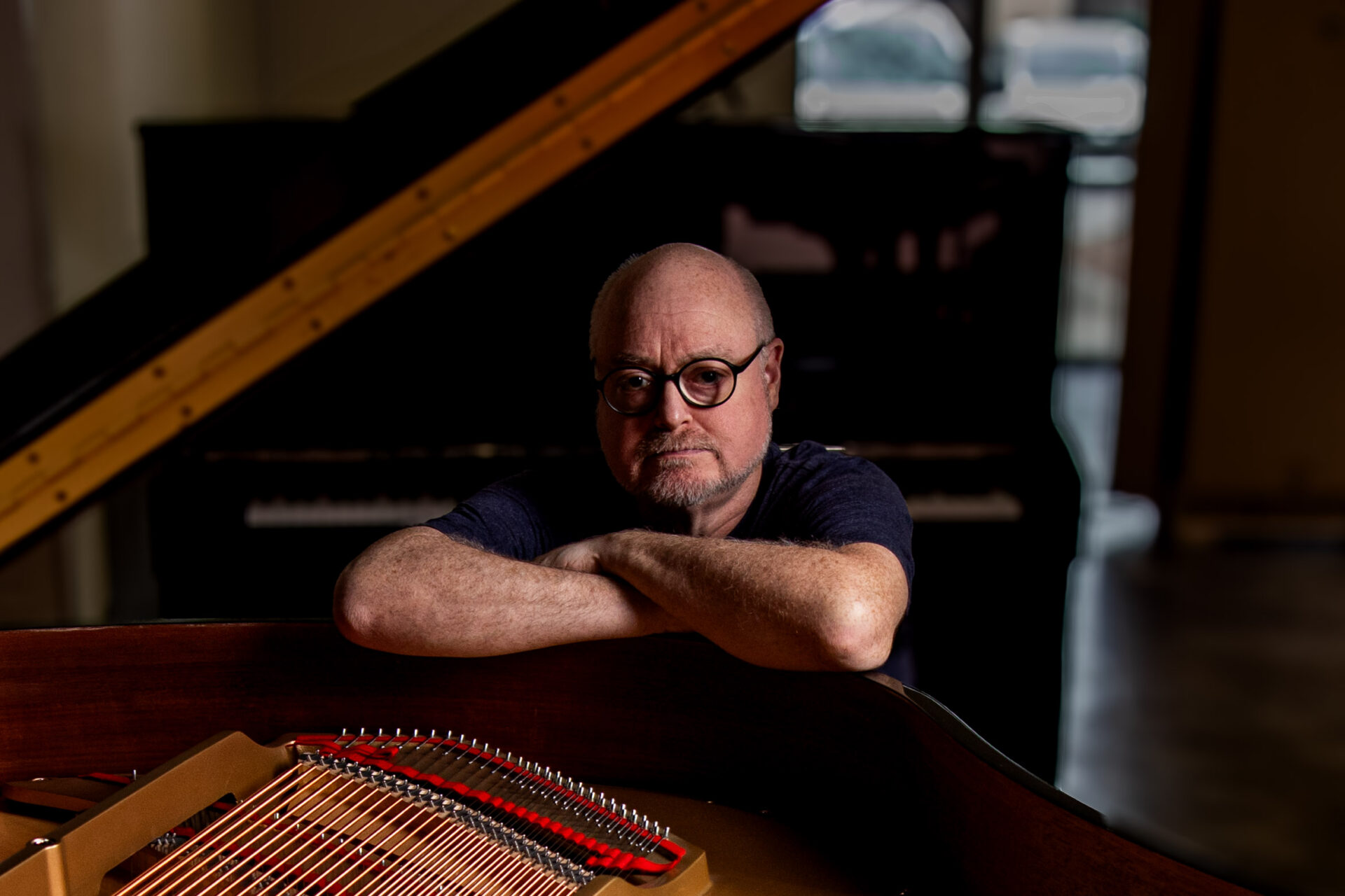 Lead Piano Technician Joel Asher rests his arms on the rim of a grand piano, with the piano’s strings visible in the foreground and another grand piano softly blurred behind him.
