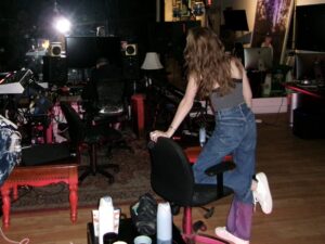 Sara Bares in a recording studio, standing beside a chair in a creative session environment with music production equipment
